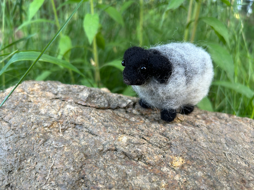 A needle-felted Icelandic sheep sits on a rock in front of tall greenery.