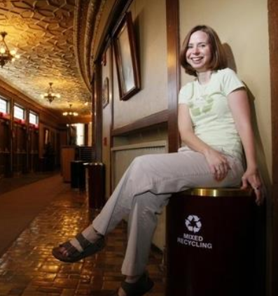 Becky sitting on a recycling can in a lobby of a theater. She is sitting cross legged with khaki pants and a green shirt with a recycle logo.