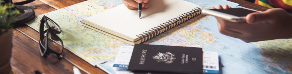 A person writes in a travel journal on top of a map, with a passport, boarding passes, camera, and glasses nearby.