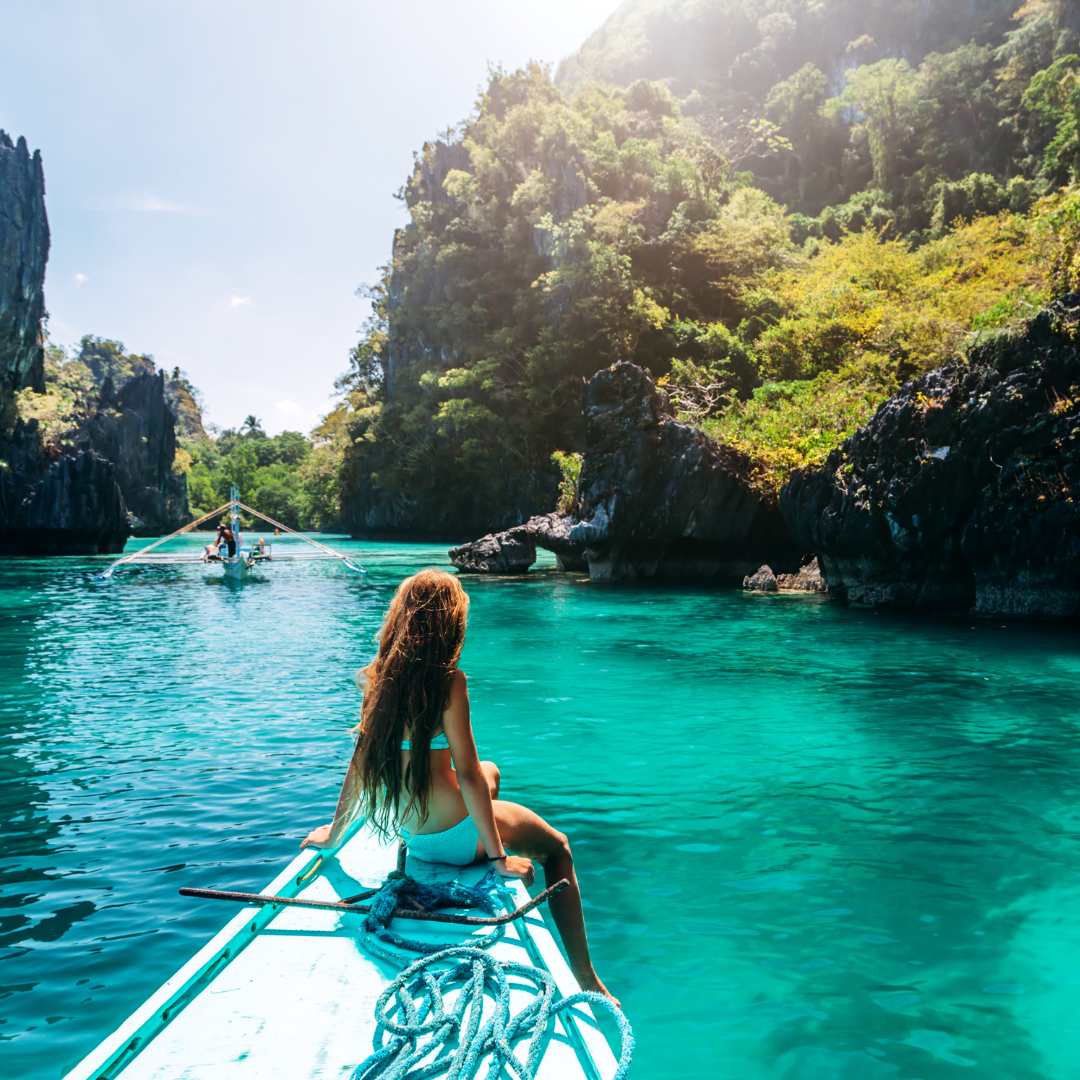 A woman sits at the front of a boat, gazing at turquoise water and lush cliffs, enjoying a peaceful travel moment.