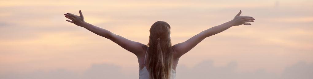A woman stands with arms outstretched, facing a sunrise or sunset, embracing the open sky.