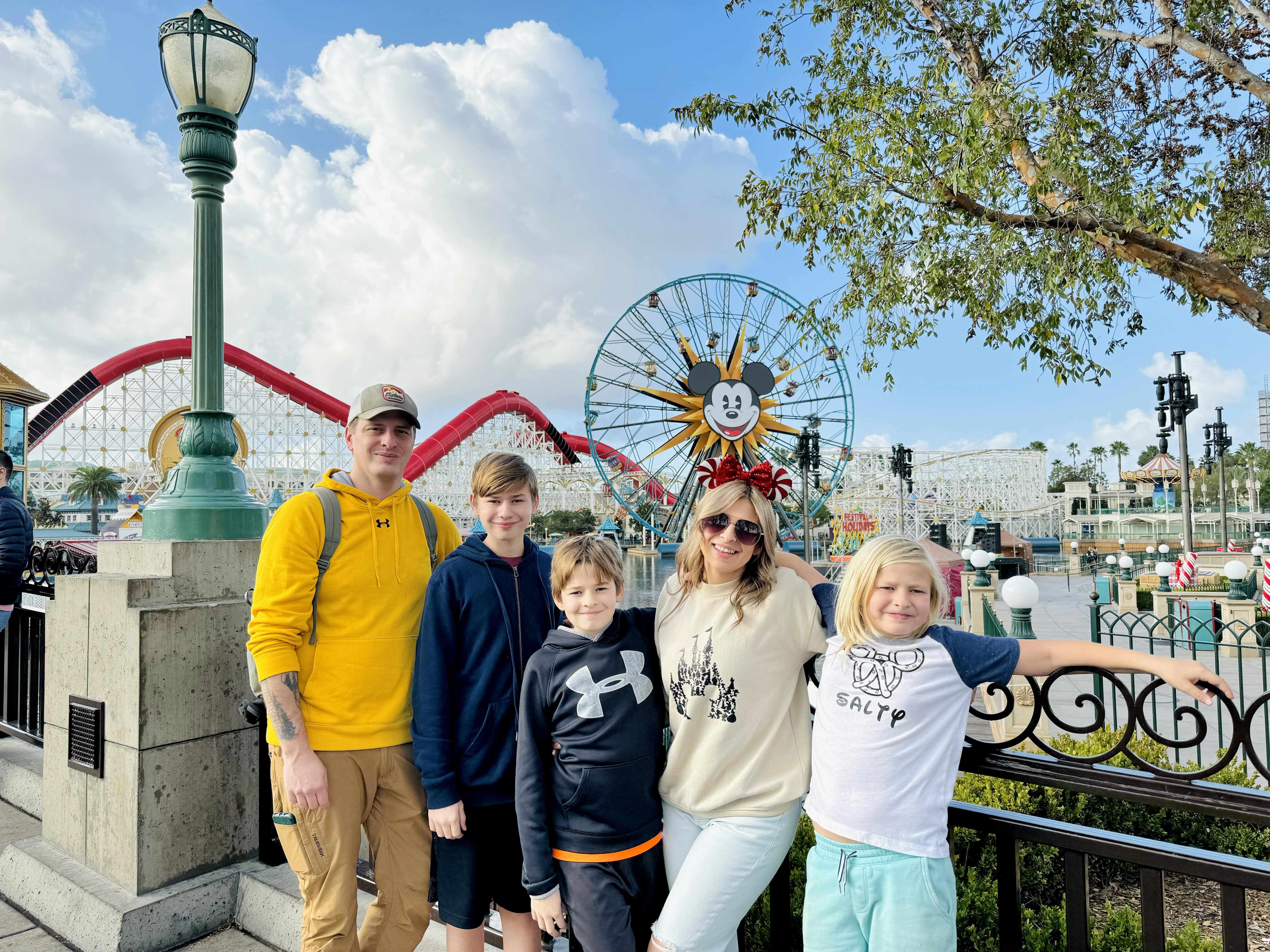 Image of a family taking their first disney vacation to disneyland resort in california. The mom is a walt disney travel agent specialzing in cruises, disney travell planning, and more. They are standing in california adveture park. Behind them is the iconic Pixar Pal-A-Round Ferris wheel and Incredicoaster. The mom, a Walt Disney World and Disneyland travel agent based in Eau Claire, Wisconsin, specializes in Disney Cruise Line vacations and planning magical Disney trips for families. They are enjoying a sunny day at the park, surrounded by festive holiday decorations.