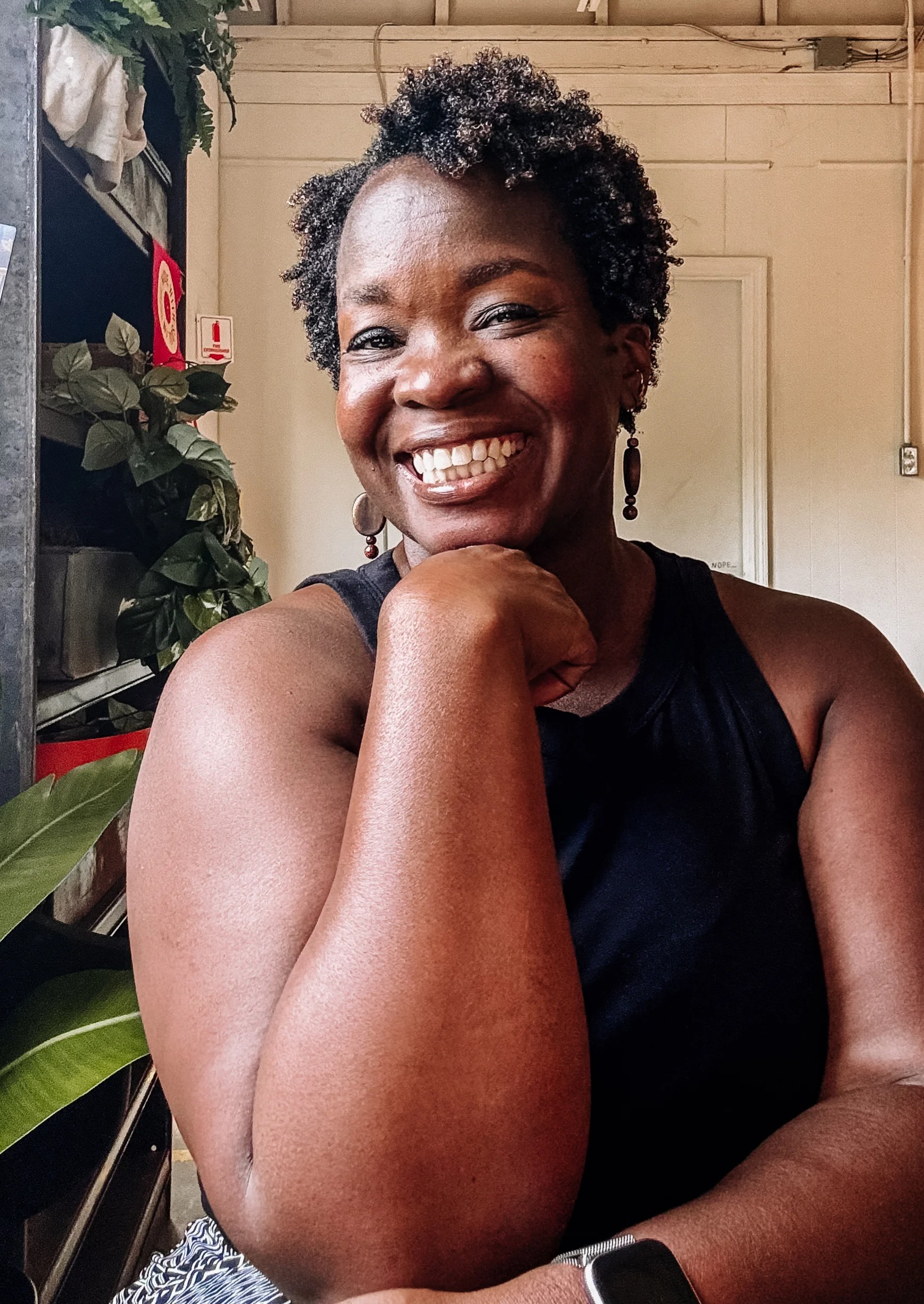 Rashidat Odeyemi smiling warmly with her chin resting on her hand, seated in front of lush green plants.