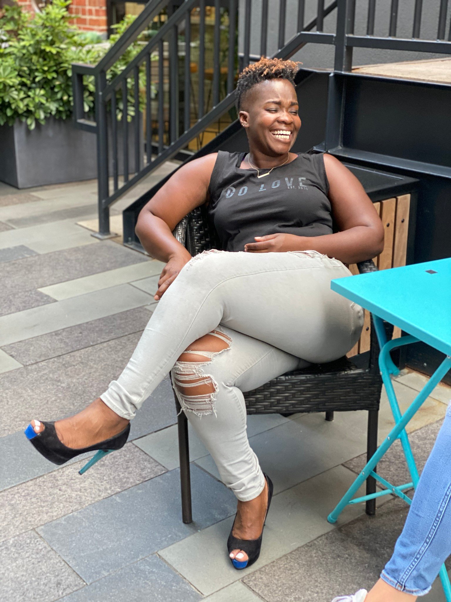 Rashidat smiling joyfully while sitting outside at a blue café table, wearing a “DO LOVE” tee and ripped jeans. Capturing the ease, authenticity, and lifestyle freedom she teaches through mindset and lifestyle coaching for ambitious Black women.