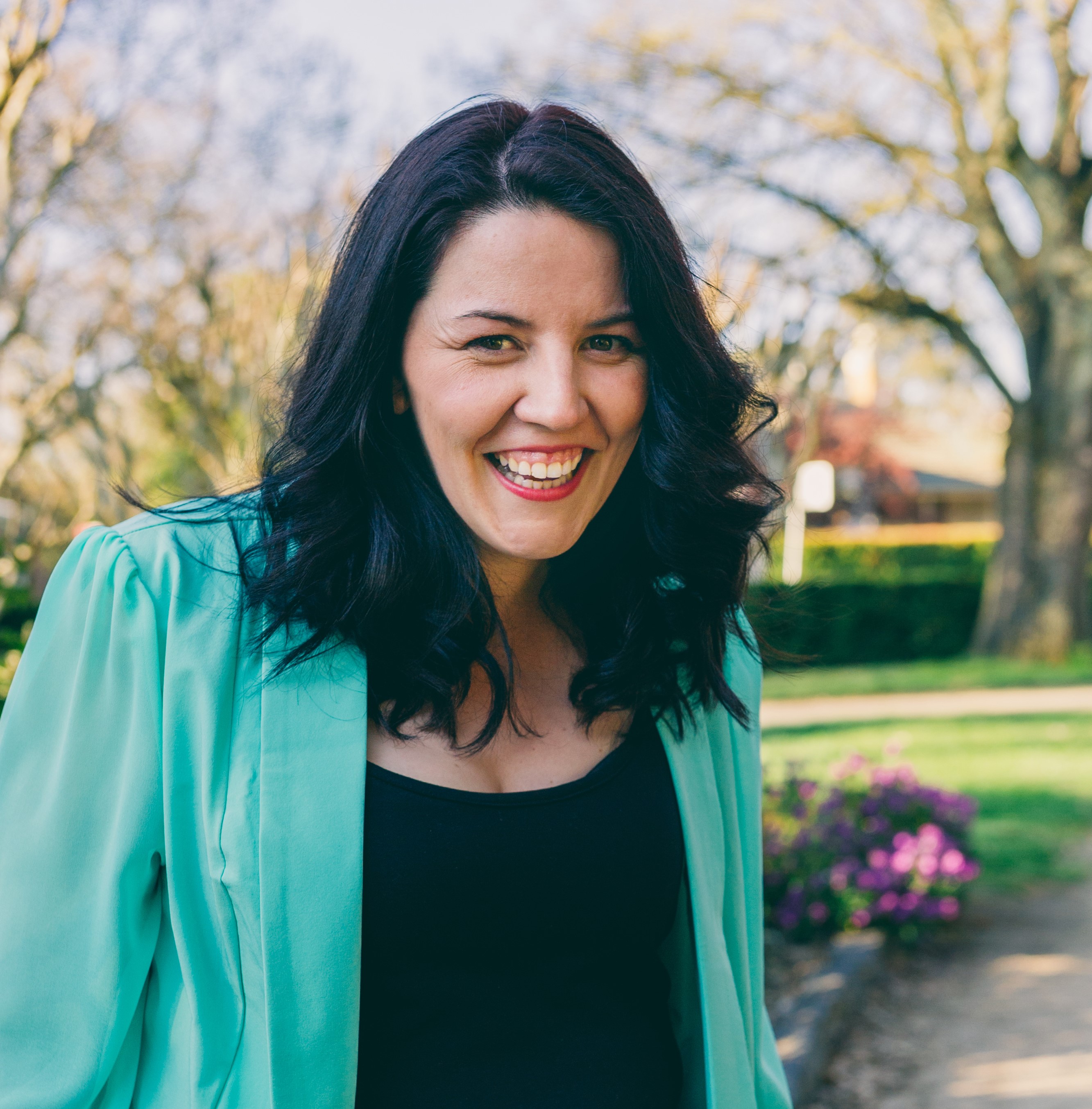 Mel Watson profile picture. A white woman with dark, shoulder length wavy hair. She is wearing a black top, aqua jacket and is smiling at the camera.