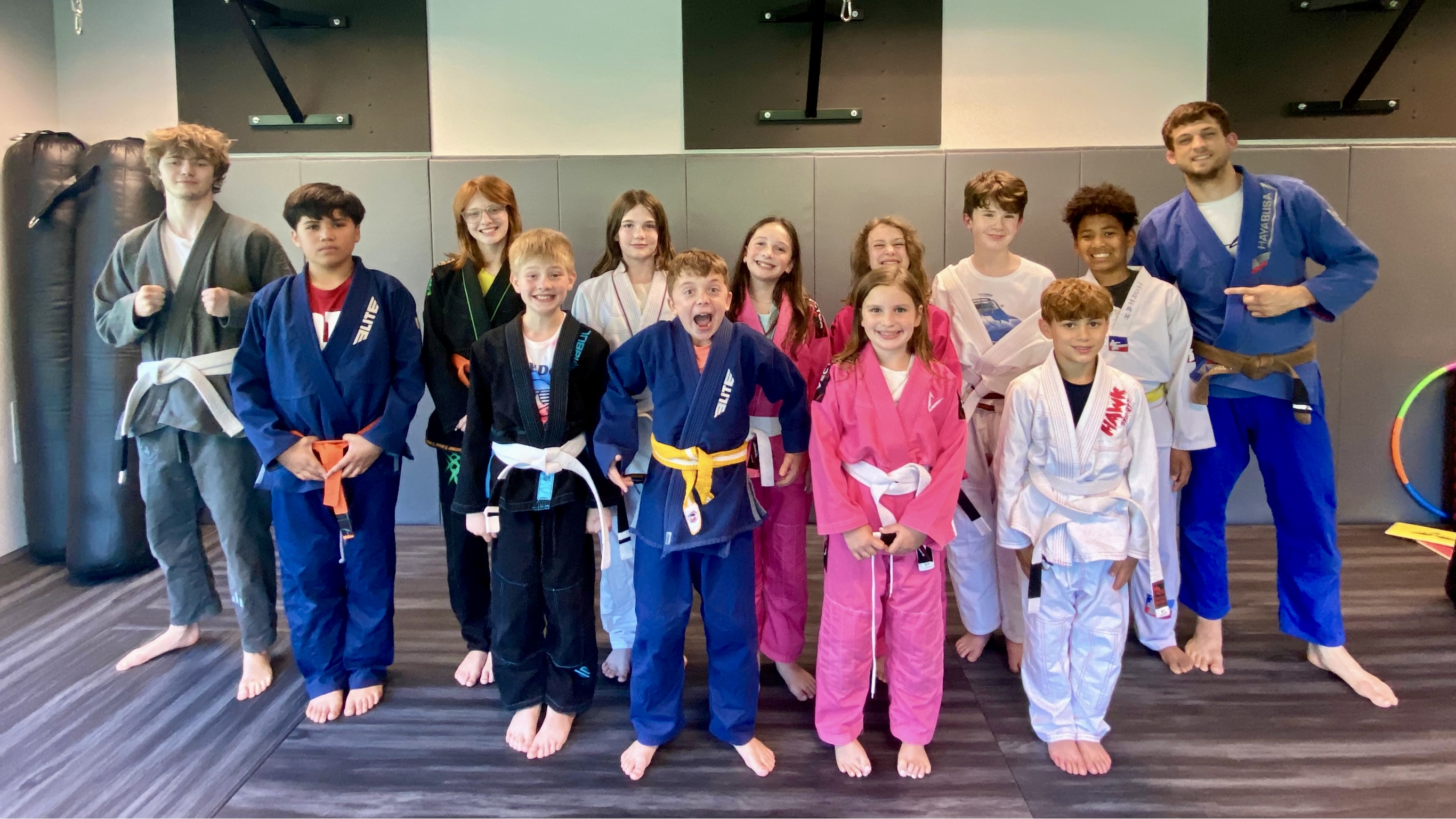 A group photo of a Brazilian Jiu-Jitsu class, featuring eleven young students and two instructors. The students are standing in two rows, wearing various colored gi (martial arts uniforms) including white, blue, pink, and black, with some wearing colored belts indicating their rank. The instructor, positioned on the far right, is dressed in a blue gi with a brown belt. The instructor positioned on the far left is dressed in a gray gi with a white belt. The group is posing on a mat in a martial arts gym with padded walls and equipment in the background, and the students have cheerful expressions.