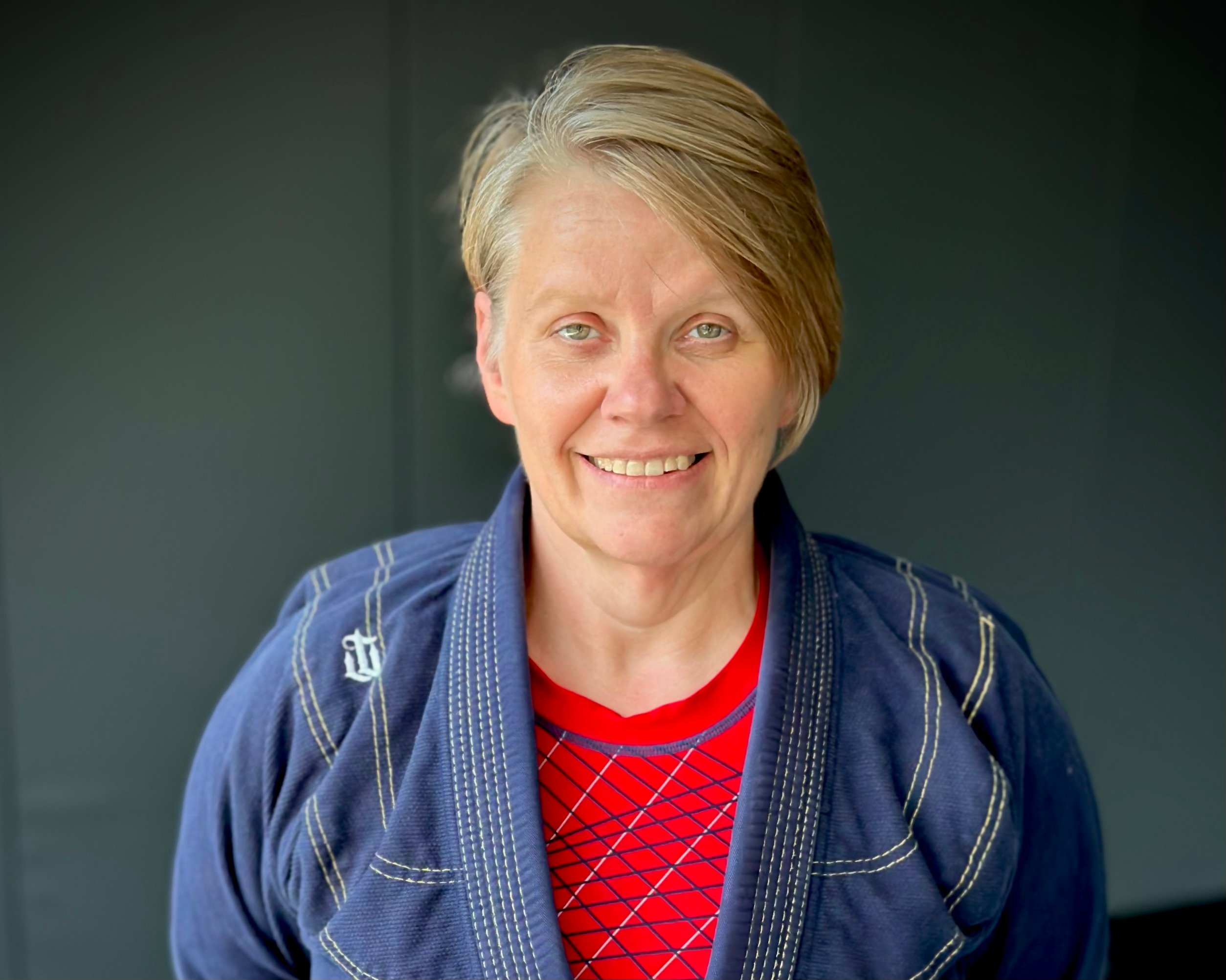 The image shows Leighia Wells wearing a blue martial arts gi with white stitching and a red shirt underneath that has a geometric pattern. She is smiling at the camera, and her short, light brown hair is neatly swept to the side. The background is a dark, neutral color, placing focus on her friendly expression and relaxed demeanor.