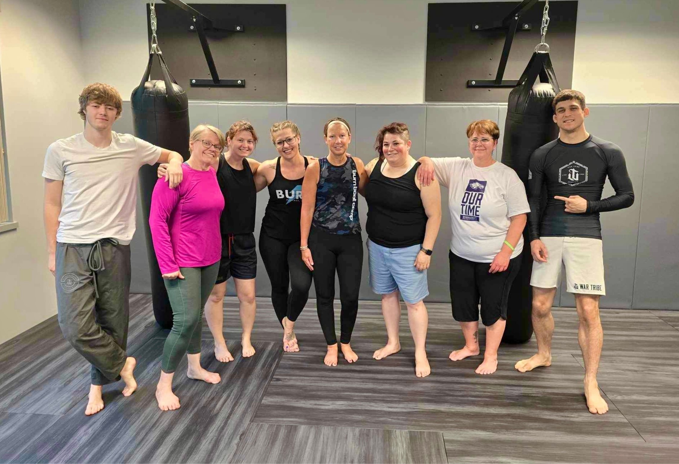 A group of eight people are standing together in a gym setting, posing for a group photo in front of two hanging punching bags. They are all barefoot and dressed in athletic attire, suggesting they have just finished or are about to start a workout session. The group consists of a mix of men and women of varying ages, all smiling and looking at the camera. The atmosphere appears to be friendly and supportive. The flooring is a dark gray, and the walls have padded sections, typical of a martial arts or fitness training area.