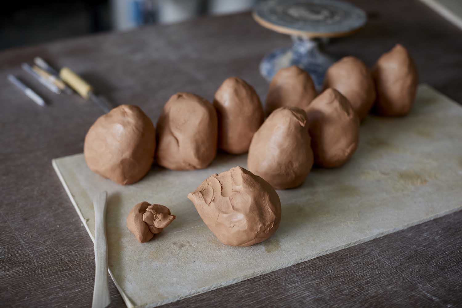 Image of a set of balls of clay in a soft brown coloured and a piece on a pale wooden board. There is a wooden tool resting on the board. In the background is a dark wooden table.