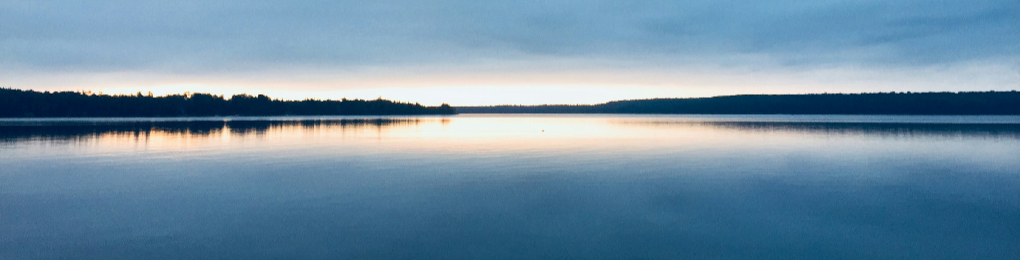 Photo of a smooth lake at sunset, with the far shoreline lined with trees in the distance. It's not clear whether the lake continues around a bend behind the line of trees.