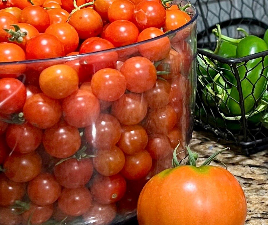 A large clear container filled with vibrant red cherry tomatoes sits on a granite countertop. In front of it, a single larger tomato with an orange tint contrasts in size and color. To the right, a black wire basket holds several glossy green jalapeño peppers.
