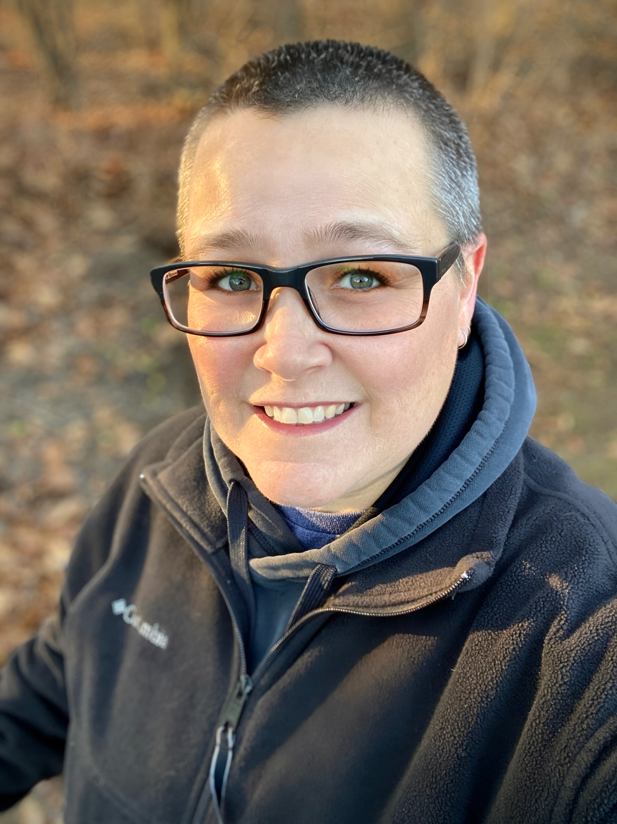 A person smiles warmly at the camera while standing in a wooded area during autumn. They wear black-framed glasses, a dark Columbia fleece jacket, and a layered hoodie. Fallen leaves cover the ground, and soft golden light—likely from early morning or late afternoon—illuminates their face. The background features tall trees with sparse foliage, suggesting a peaceful, crisp outdoor setting.