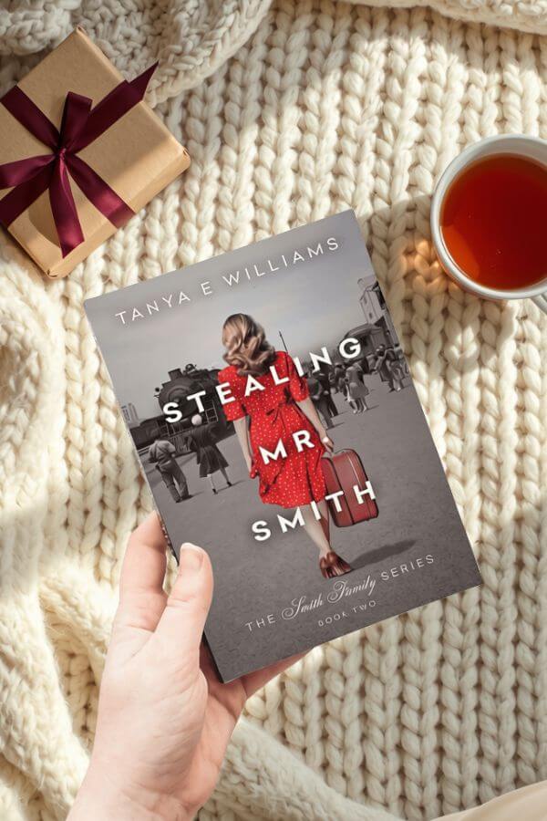 A hand holding Stealing Mr. Smith by Tanya E Williams, styled with a wrapped gift and a cup of tea for a holiday reading gift guide.