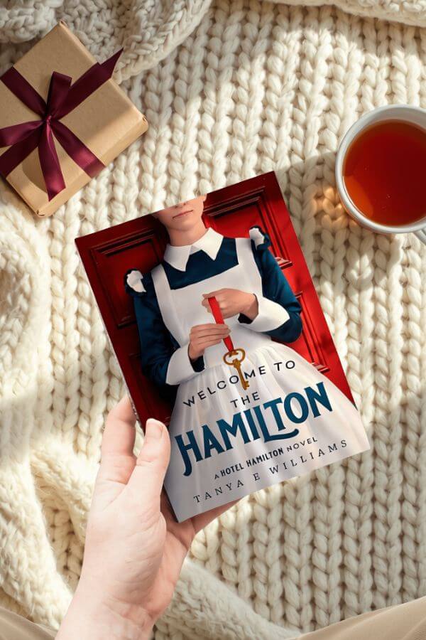 A hand holding Welcome to the Hamilton by Tanya E Williams, styled with a wrapped gift and a cup of tea for a holiday reading gift guide.