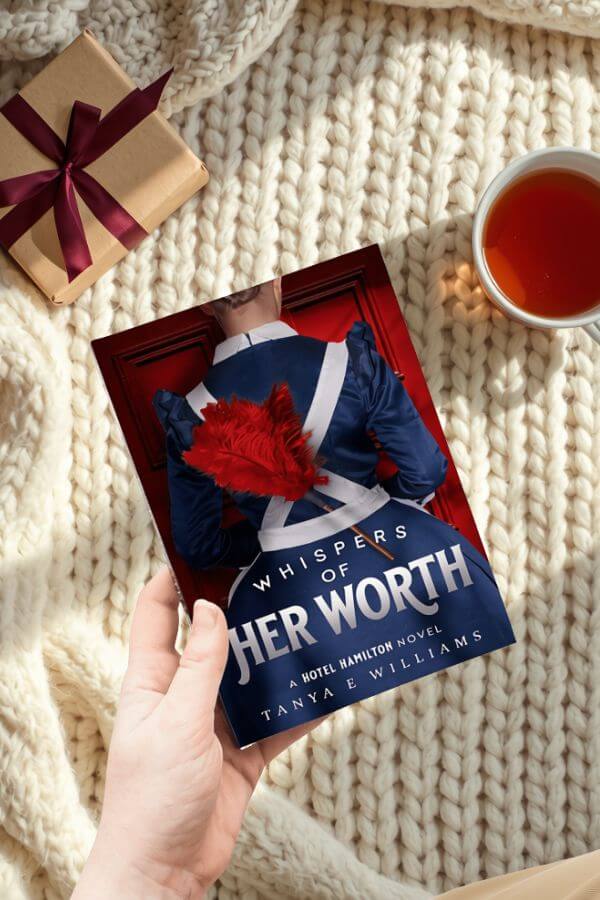 A hand holding Whispers of Her Worth by Tanya E Williams, styled with a wrapped gift and a cup of tea for a holiday reading gift guide.