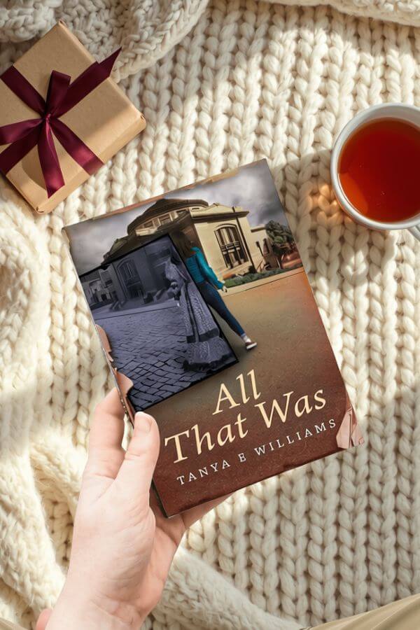 A hand holding All That Was by Tanya E Williams, styled with a wrapped gift and a cup of tea for a holiday reading gift guide.