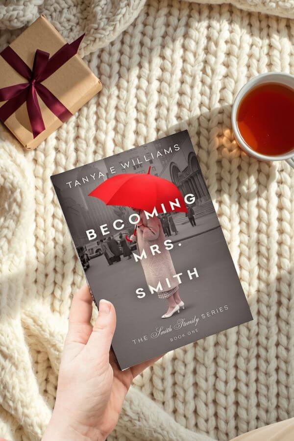 A hand holding Becoming Mrs. Smith by Tanya E Williams, styled with a wrapped gift and a cup of tea for a holiday reading gift guide.