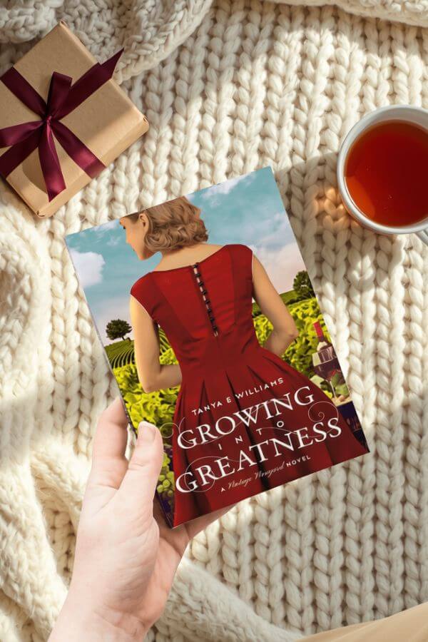 A hand holding Growing into Greatness by Tanya E Williams, styled with a wrapped gift and a cup of tea for a holiday reading gift guide.