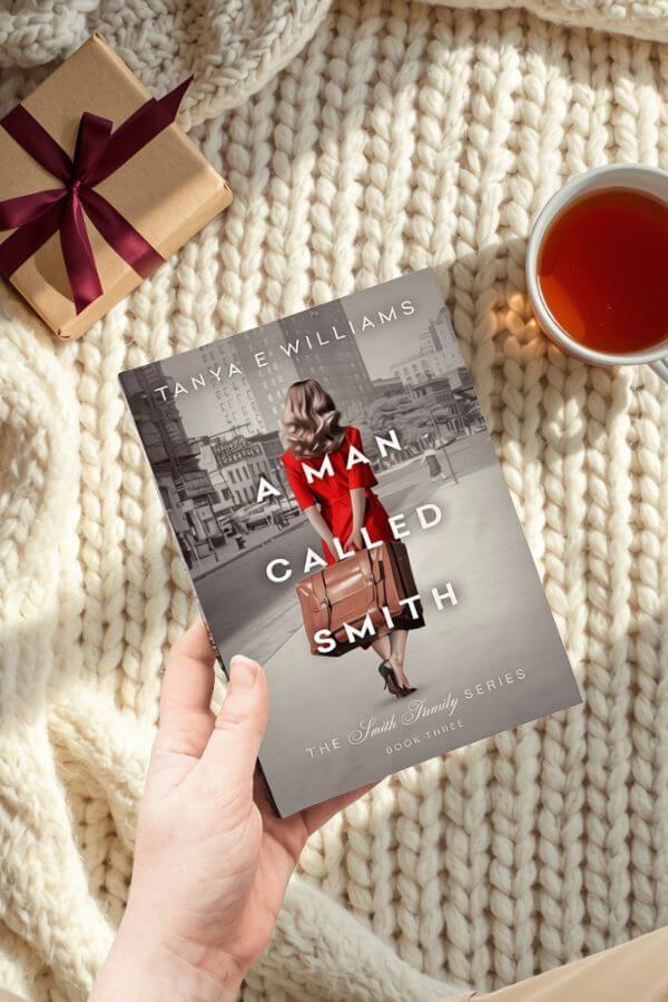 A hand holding A Man Called Smith by Tanya E Williams, styled with a wrapped gift and a cup of tea for a holiday reading gift guide.