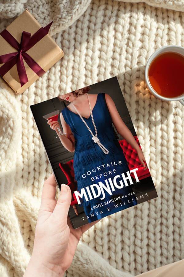A hand holding Cocktails Before Midnight by Tanya E Williams, styled with a wrapped gift and a cup of tea for a holiday reading gift guide.