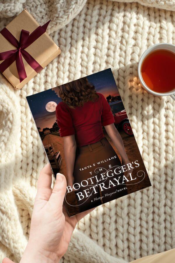 A hand holding The Bootlegger’s Betrayal by Tanya E Williams, styled with a wrapped gift and a cup of tea for a holiday reading gift guide.
