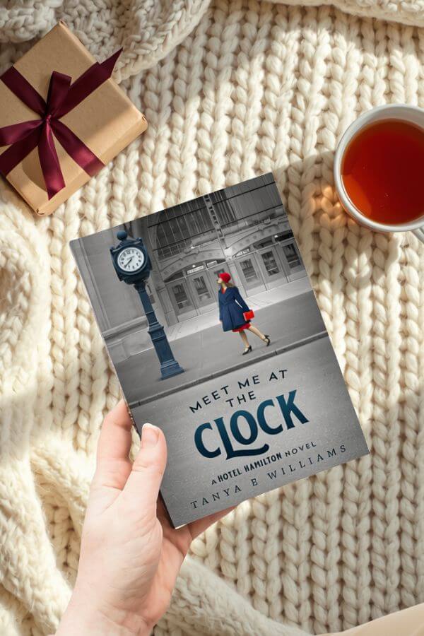 A hand holding Meet Me at the Clock by Tanya E Williams, styled with a wrapped gift and a cup of tea for a holiday reading gift guide.