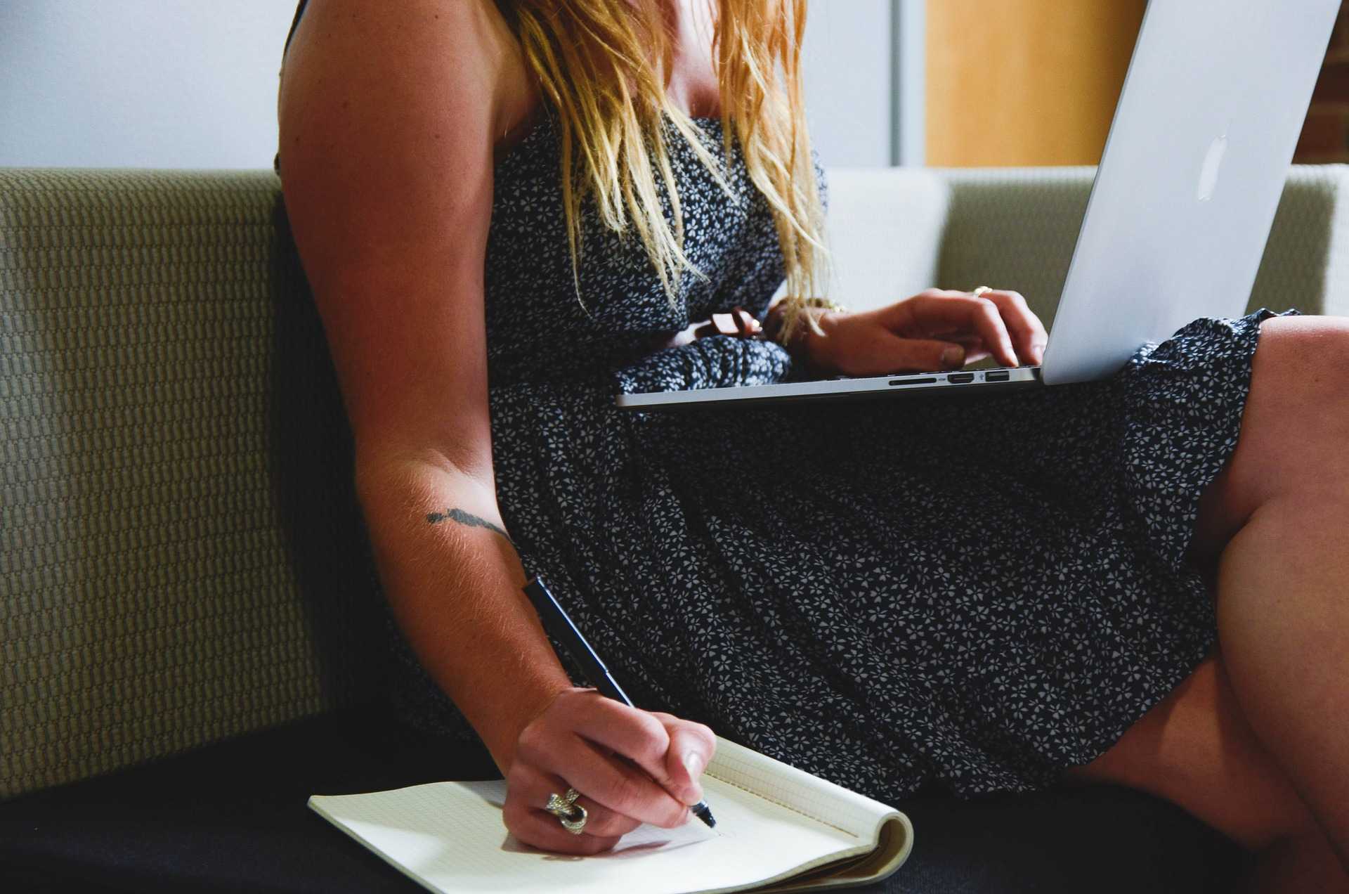 Image of a mid-sized white feminine-appearing person from the shoulders down, with a laptop on their lap, taking notes during their online therapy session