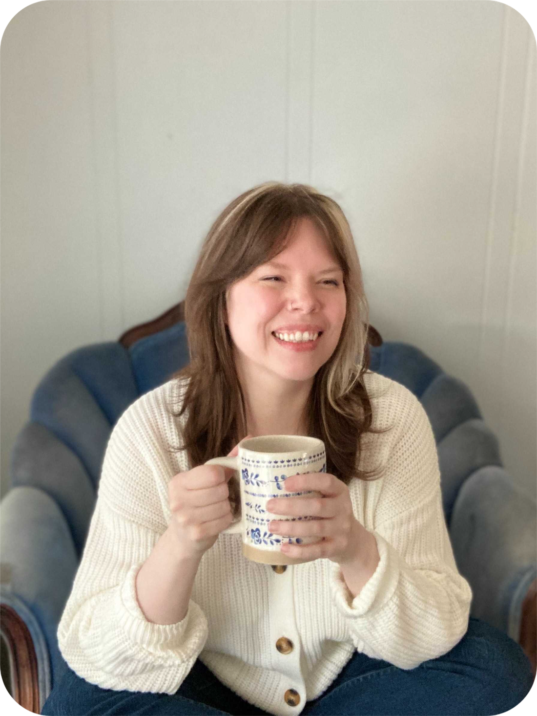 An image of Brandy, a mid-sized white woman, sitting on a blue chair, holding a cup of tea and smiling