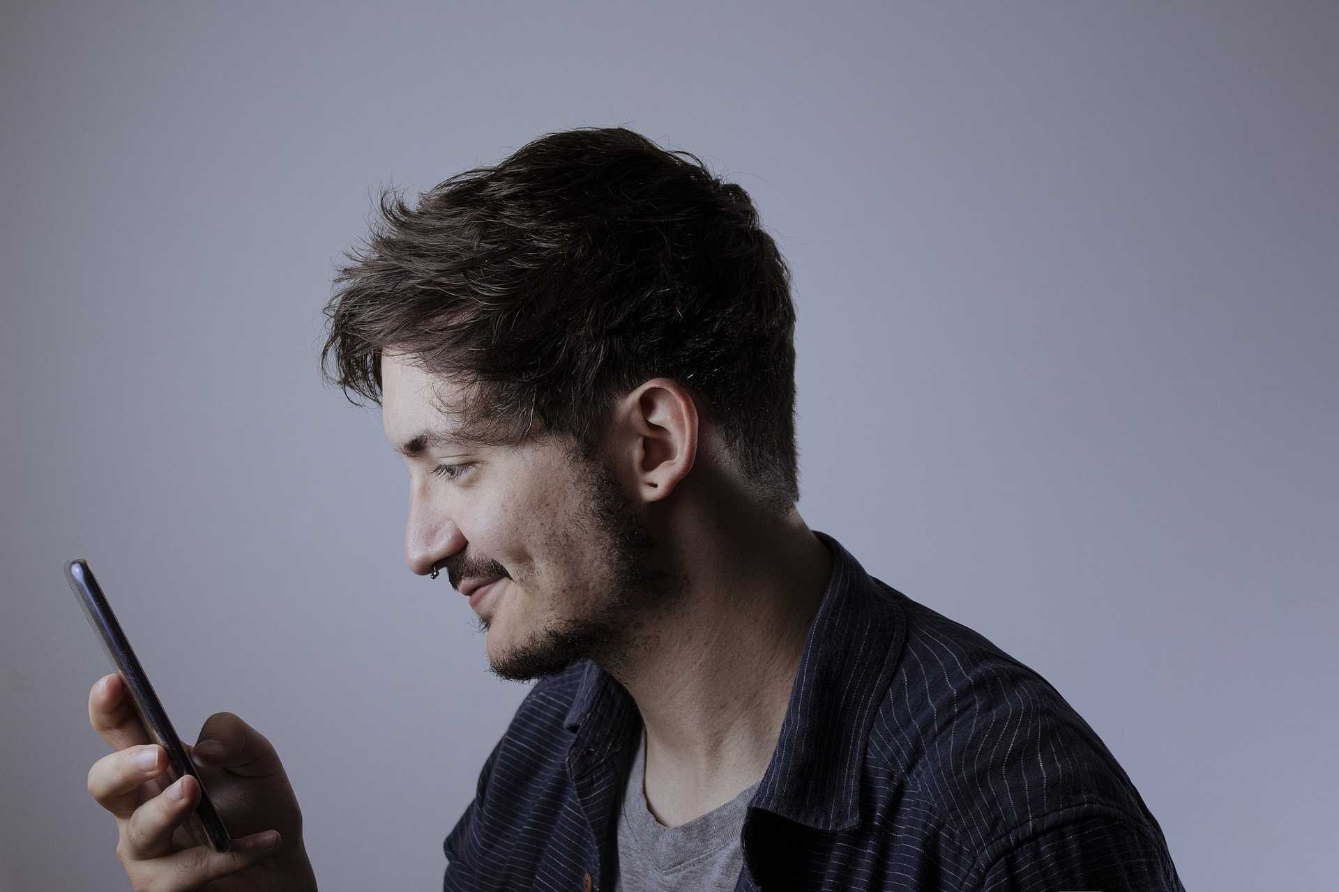 Image of a white masculine figure with a septum piercing, smiling into their phone during telehealth therapy