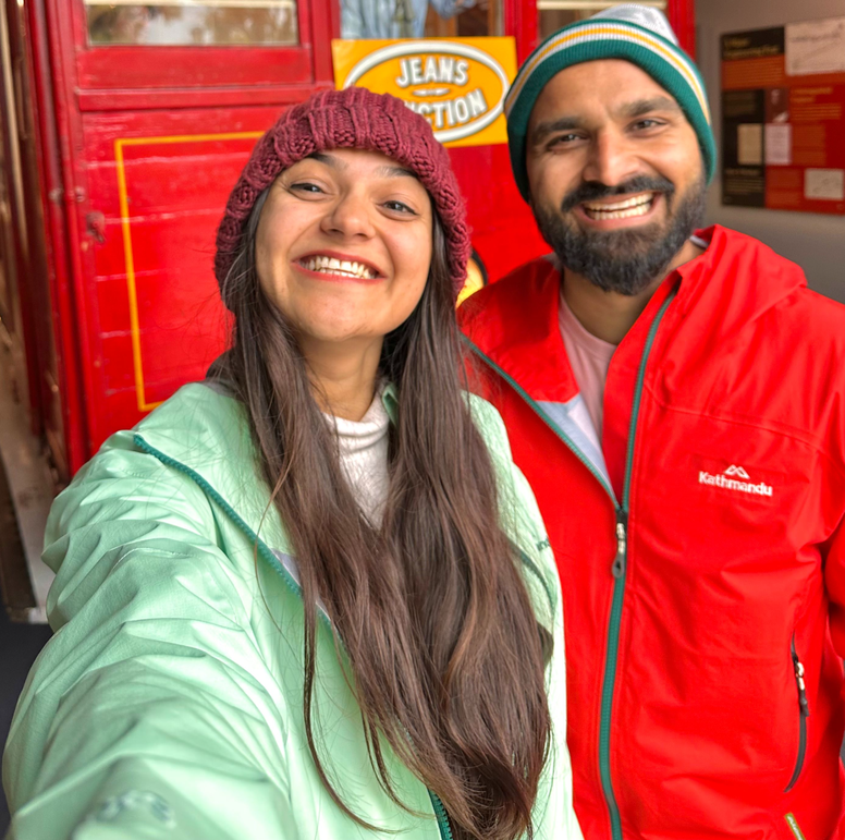 A smiling couple wearing matching pink traditional ponchos poses with two elephants at an ethical elephant sanctuary. The elephants stand calmly in a natural setting with lush green trees and a cloudy sky in the background. The couple looks happy, enjoying their wildlife experience.