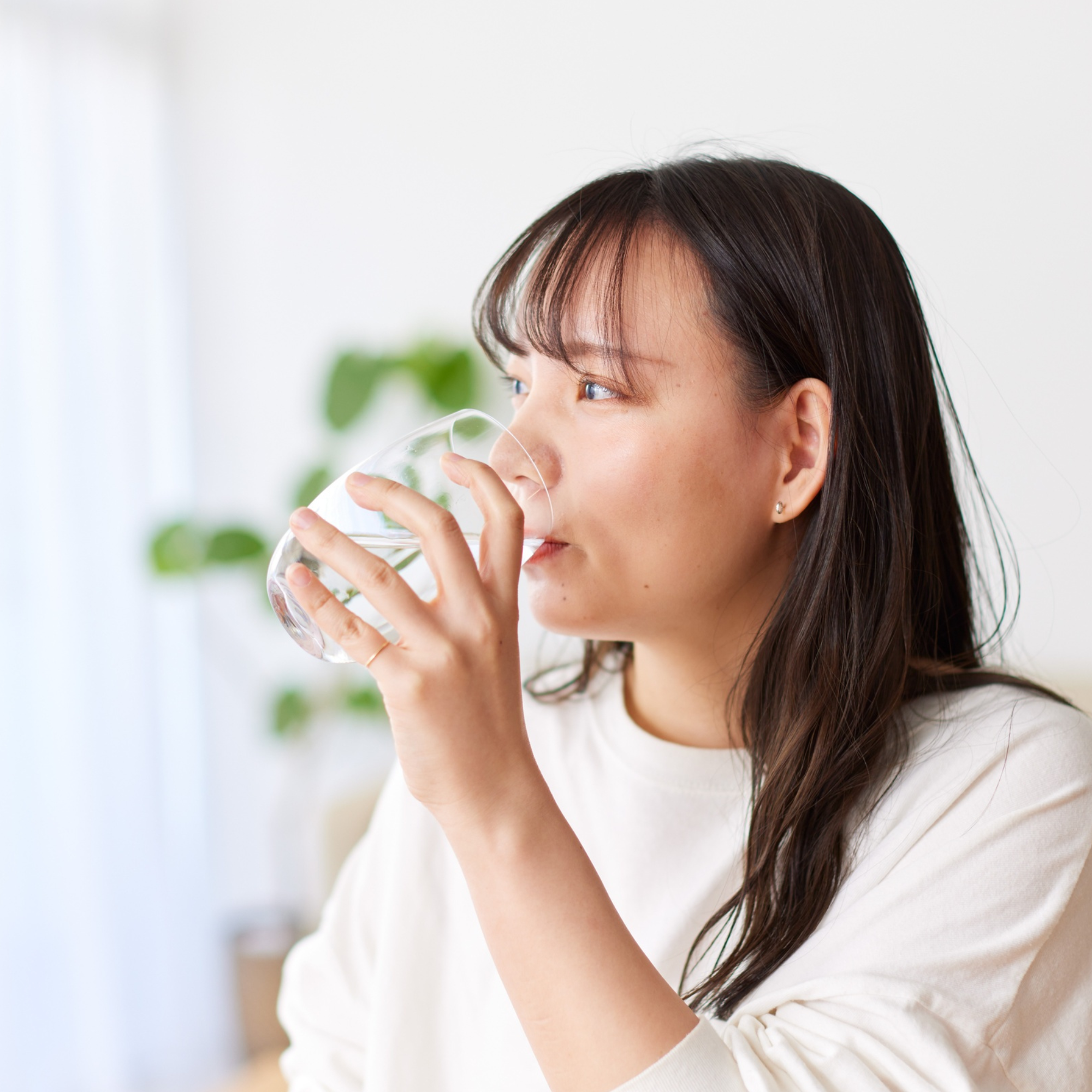 Young woman drinking a glass of water indoors, promoting hydration and vocal health in a calm, bright home setting.