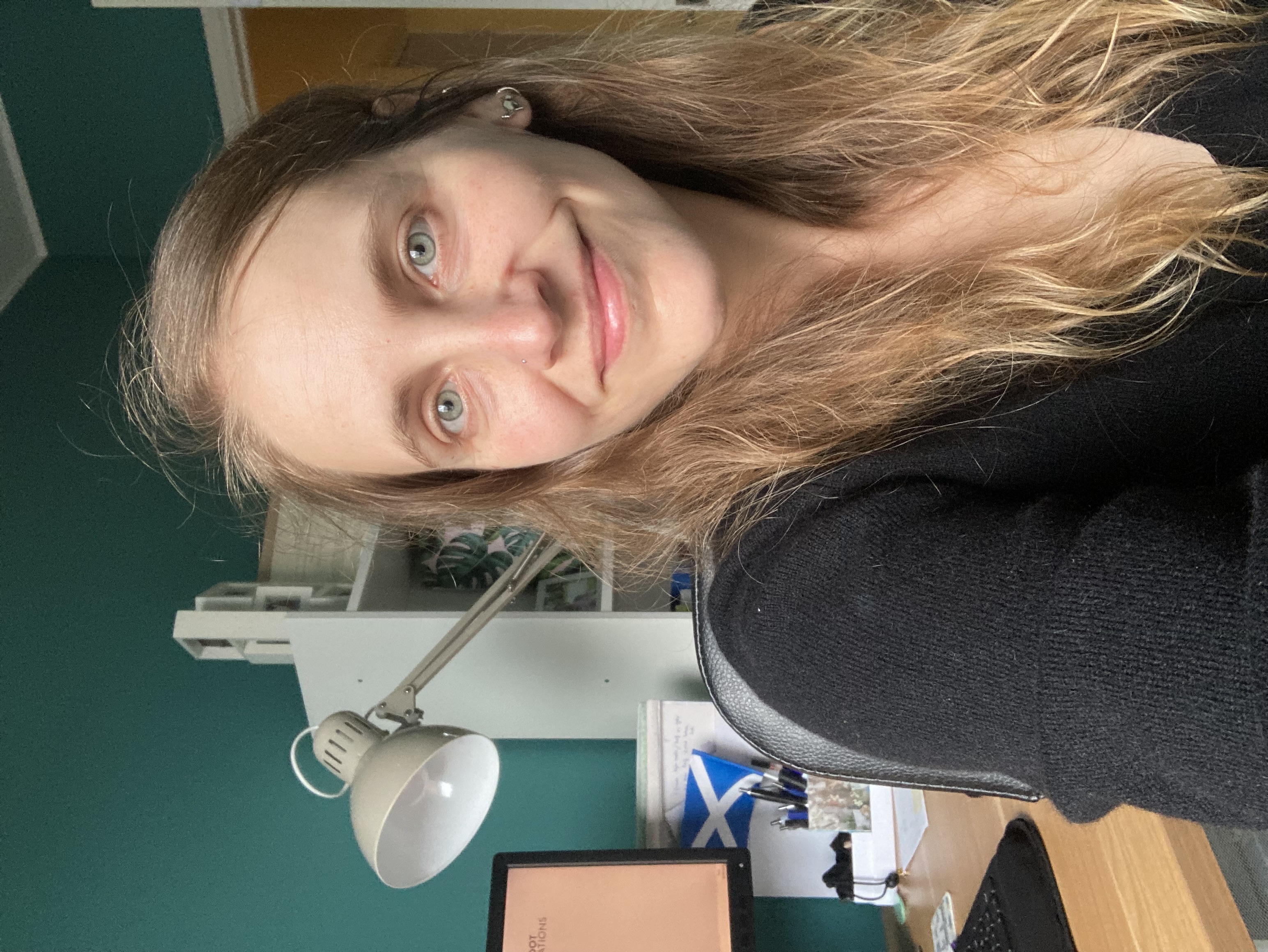 Alicja, a white woman in her 30's with dark blond wavy hair, sits are her desk. She looks at the camera smiling.