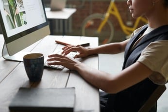 A woman sitting at a desk, writing her actual grant proposal rather than a 5-page narrative CV. Some notebooks and a hot beverage are also on the desk.