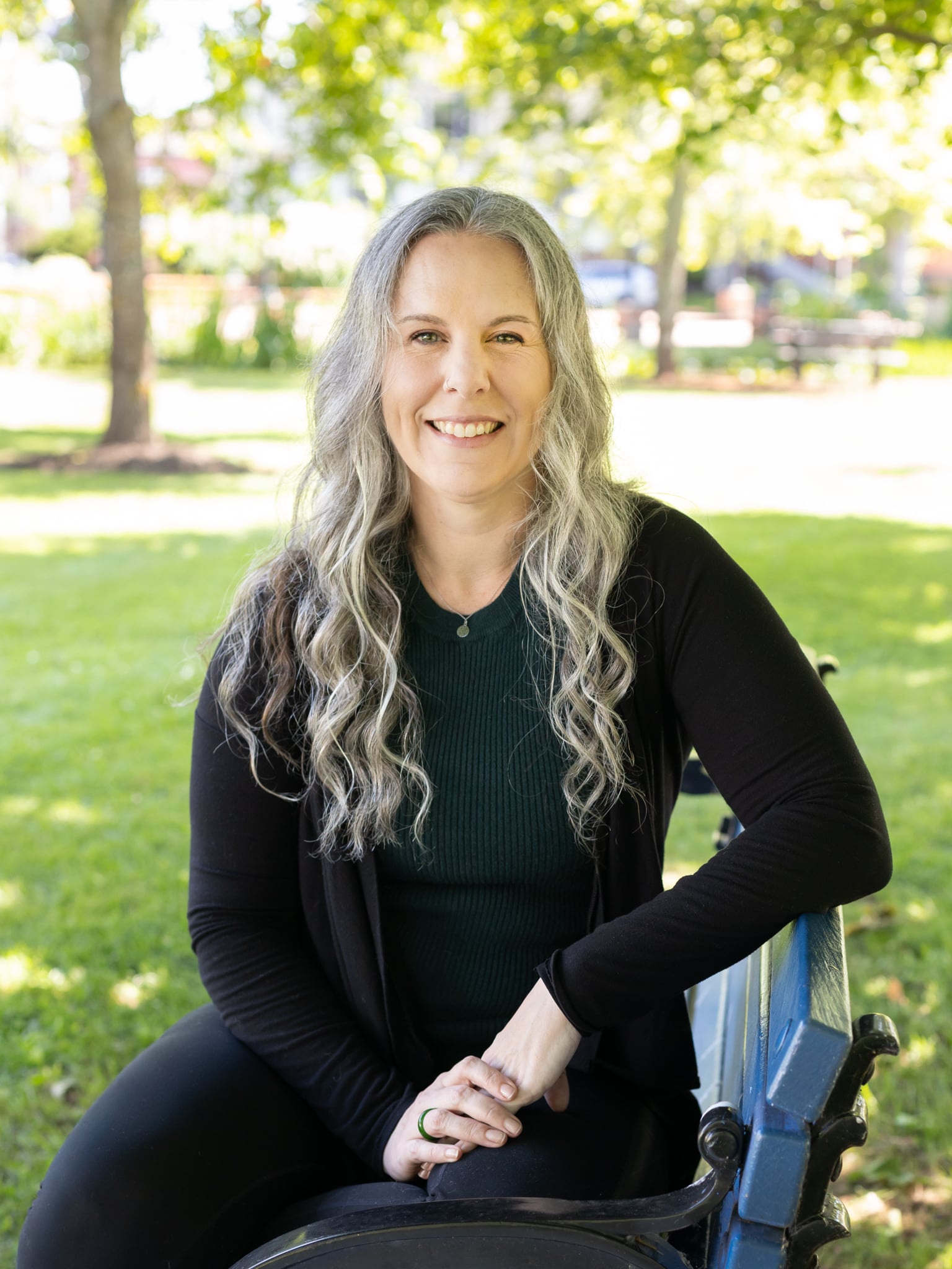 A photo of High-Fidelity's founder Dr. Nicole St. Denis, leaning against a grey exterior wall and trying to look professional and not at all uncomfortable taking business headshots. She is a white woman with long grey curly hair and green eyes, wearing a teal dress and a black blazer. Behind her is a body of water with an island in the distance.