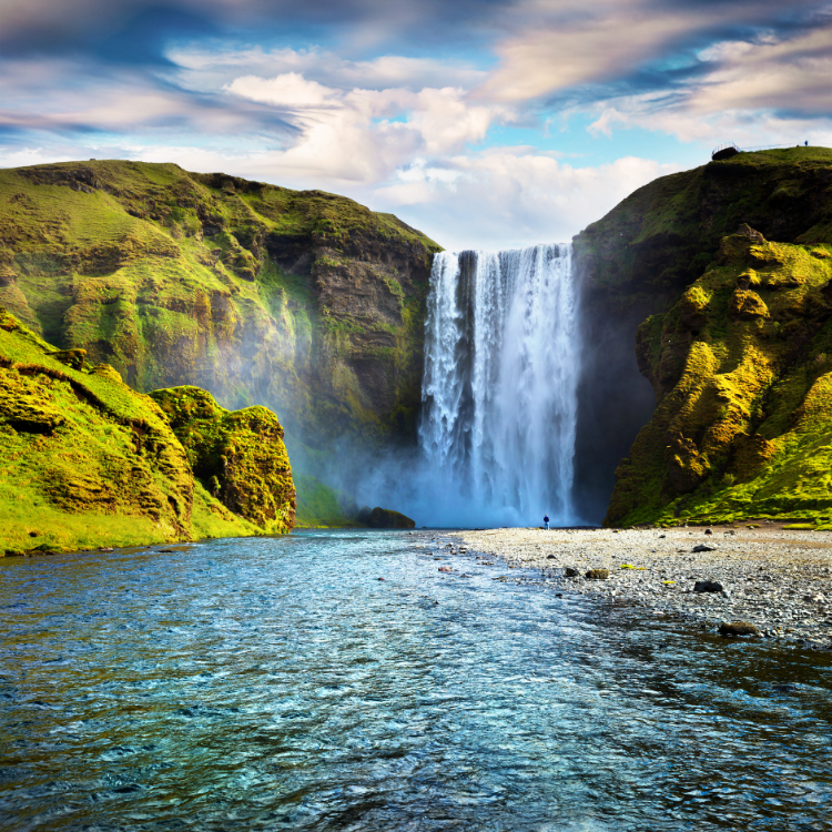 A powerful waterfall cascades down a moss-covered cliff into a clear blue river, surrounded by lush green hills under a dramatic sky.