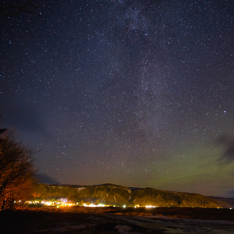 A vast star-filled night sky stretches over a quiet, illuminated town nestled at the base of dark, snow-dusted mountains.