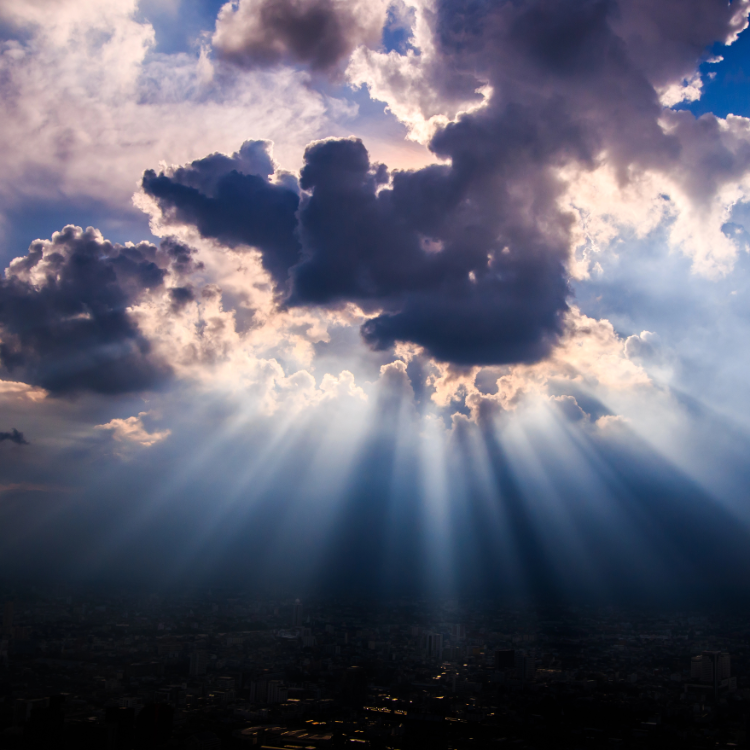 Dramatic sunbeams pierce through thick, dark storm clouds, casting light over a cityscape shrouded in shadow below.