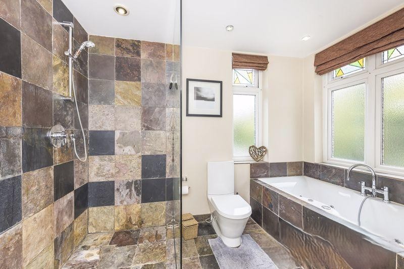 Family bathroom with indian slate tiles to walls and floor, floor to ceiling glass panel next to wet room shower area. Jacuzzi bath and WC with windows on two elevations.