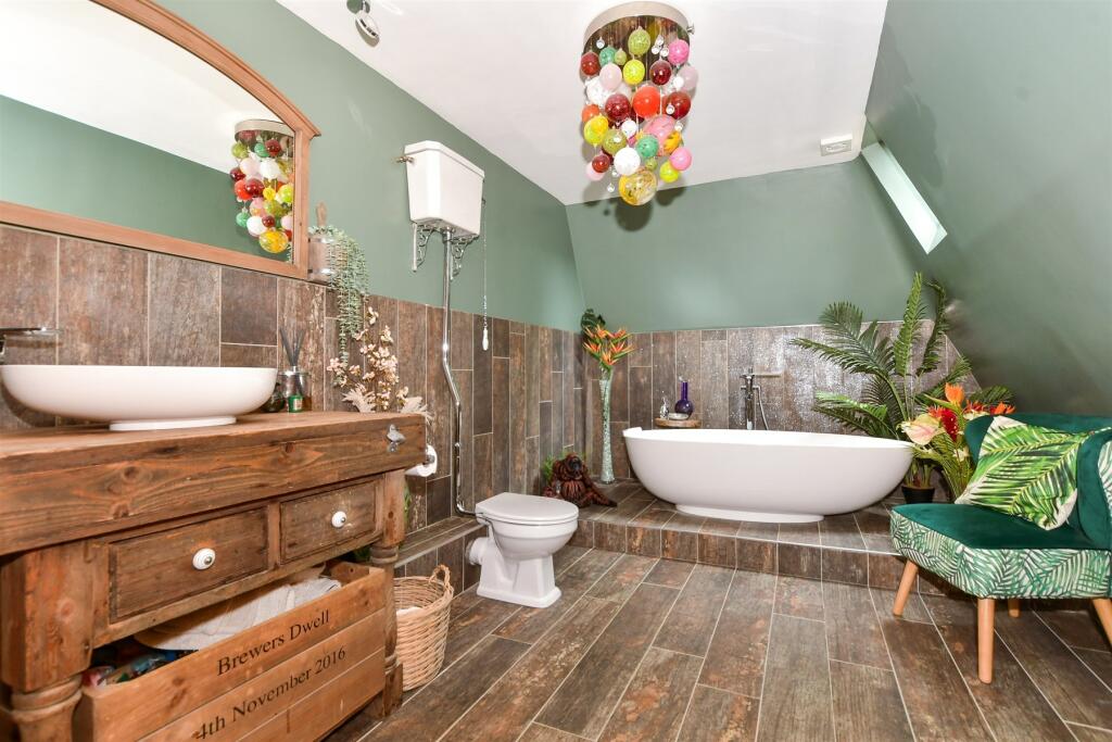 Bathroom with free standing antique sink stand in oak with white porcelain basin, mirror over the basin, towels in bespoke crate below unit. Victorian style WC with chain pull and step up on wood effect tiled floor to free standing bath in white ceramic with freestanding taps. Green walls above tiles and green velvet chair with leaf effect to the side and ceiling light with multi colour glass baubles. Sanitary ware fitted by CDI Contractors