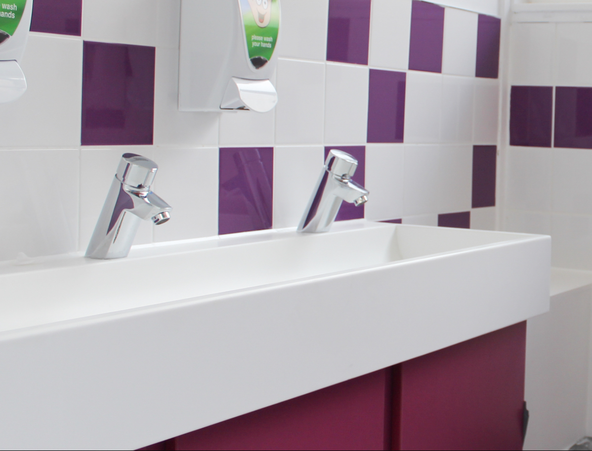 School WC Toilet Area showing white trough basin with stainless steel tap and white soap dispenser on purple and white square tiled wall in a purple base unit