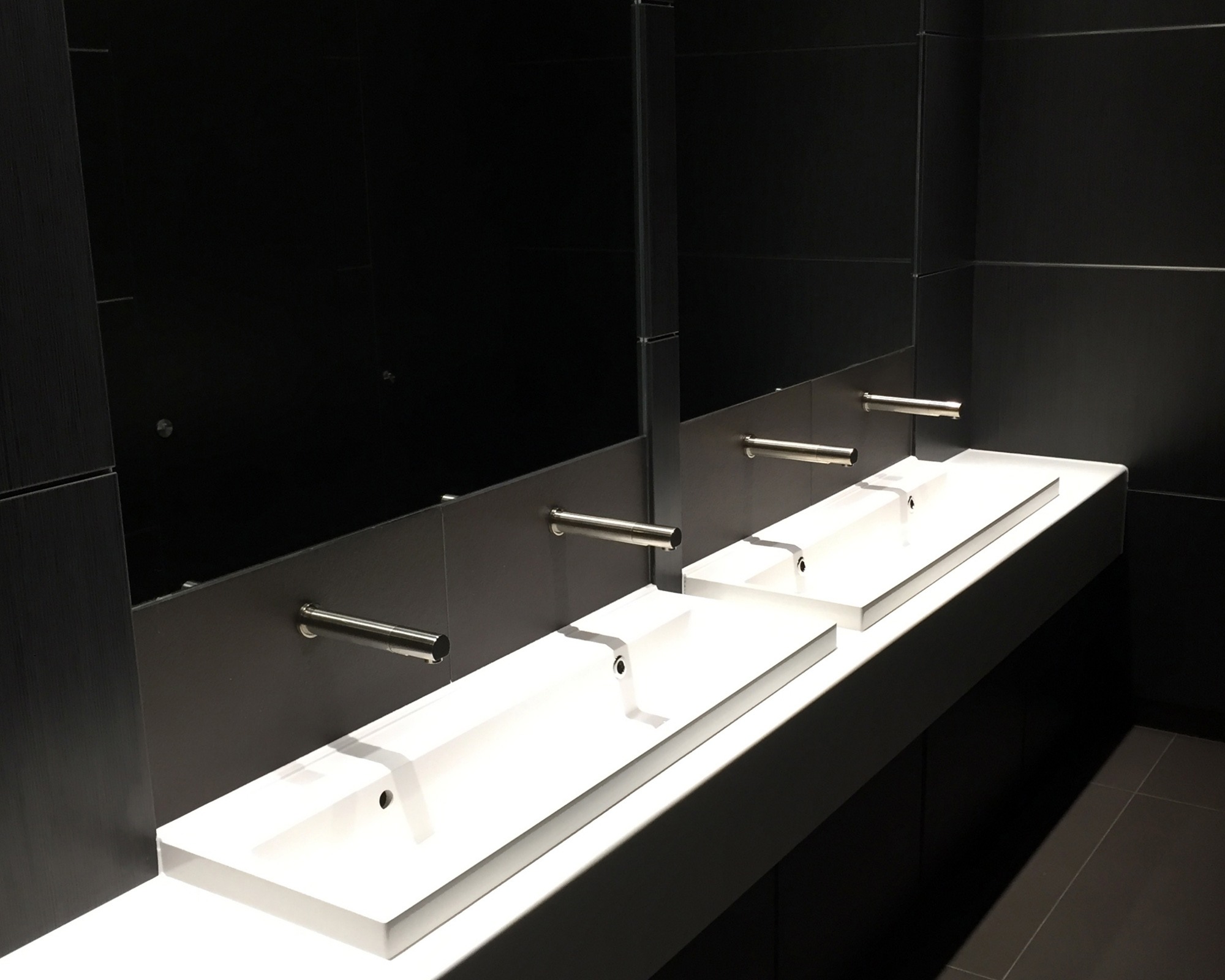 Black ash duct panel splash back in an office space WC Toilet area with white trough sinks and wall mounted stainless steel sink units in black ash base unit