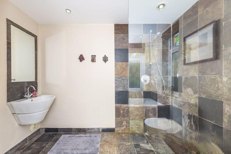 Family bathroom with indian slate tiles to walls and floor, floor to ceiling glass panel next to wet room shower area. white conical shaped basin with stainless steel mixer tap
