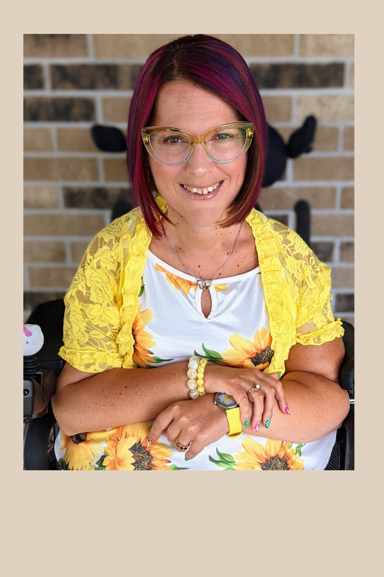 Photo of Erica Andres. Middle aged woman with red purple har. Wearing glasses. Sitting in a wheelchair in front of a brick wall.