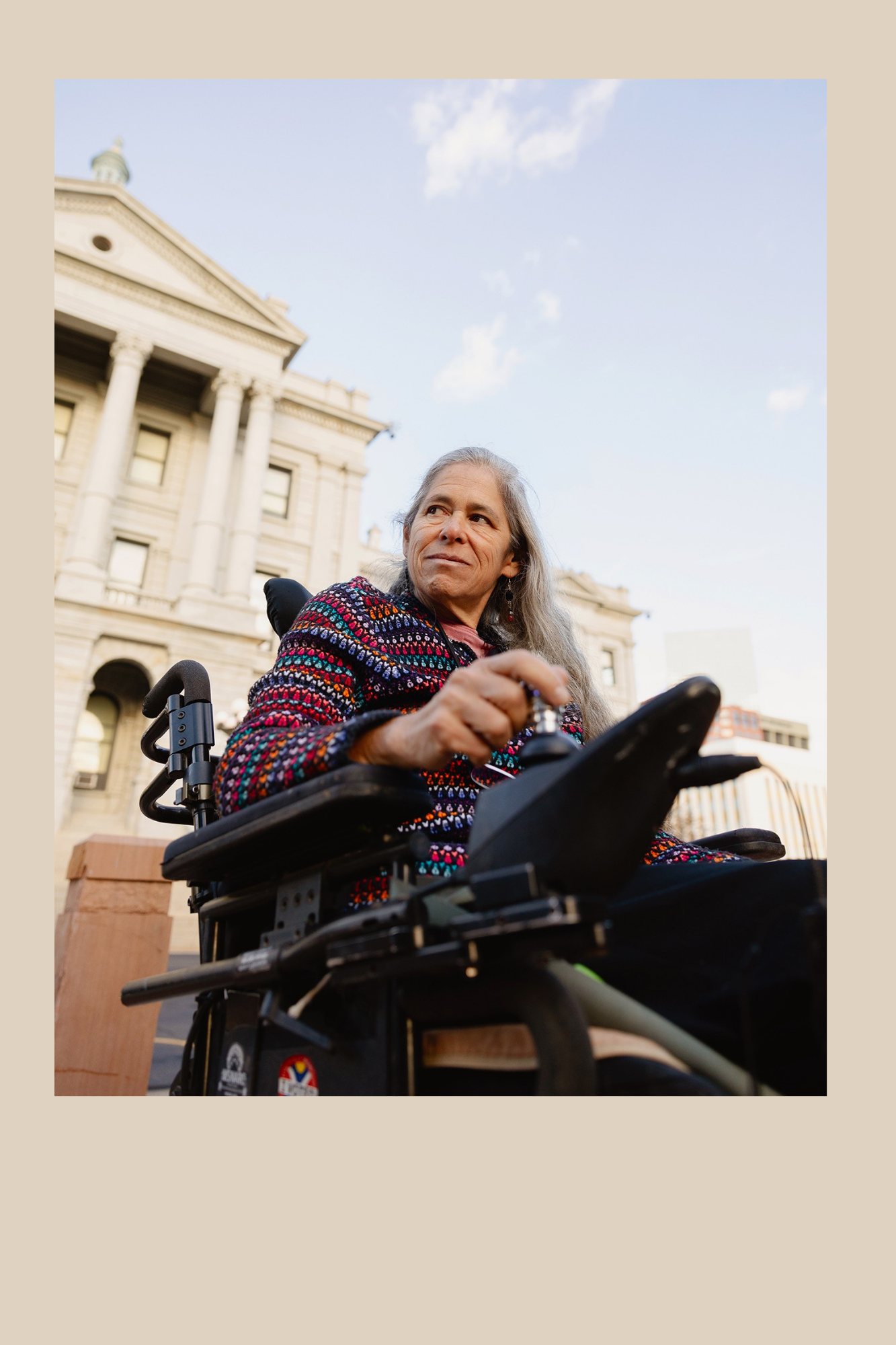 Photo of Julie Reiskin. Shi is a middle aged woman with long grey hair. She is sitting in a wheelchair in front of a state capital.