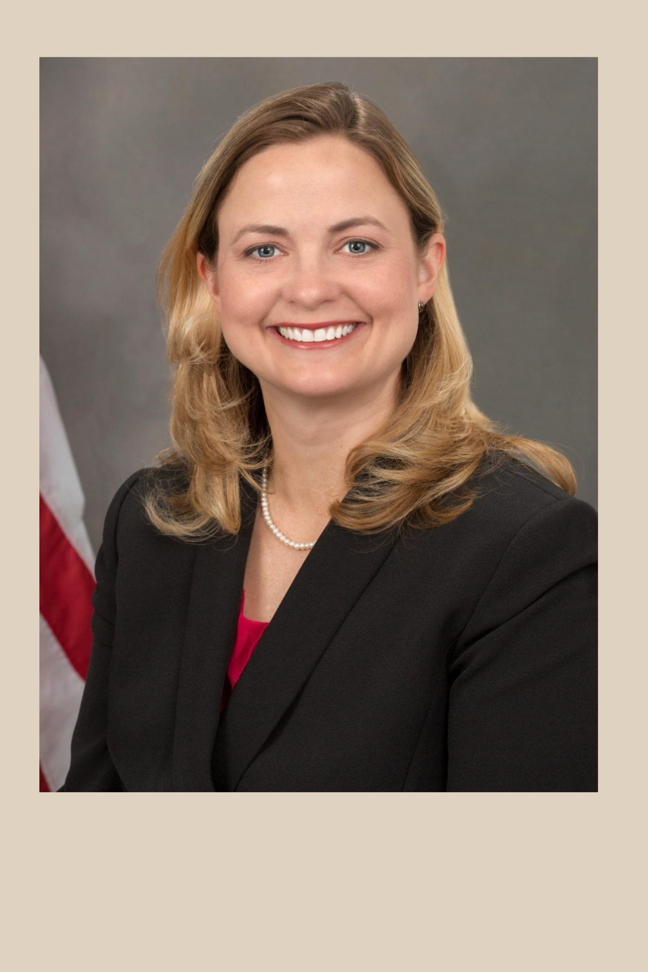 Headshot of Serena Lowe. She is standing near an American flag. She is smiling. She has long blond hair and is wearing a black blazer.