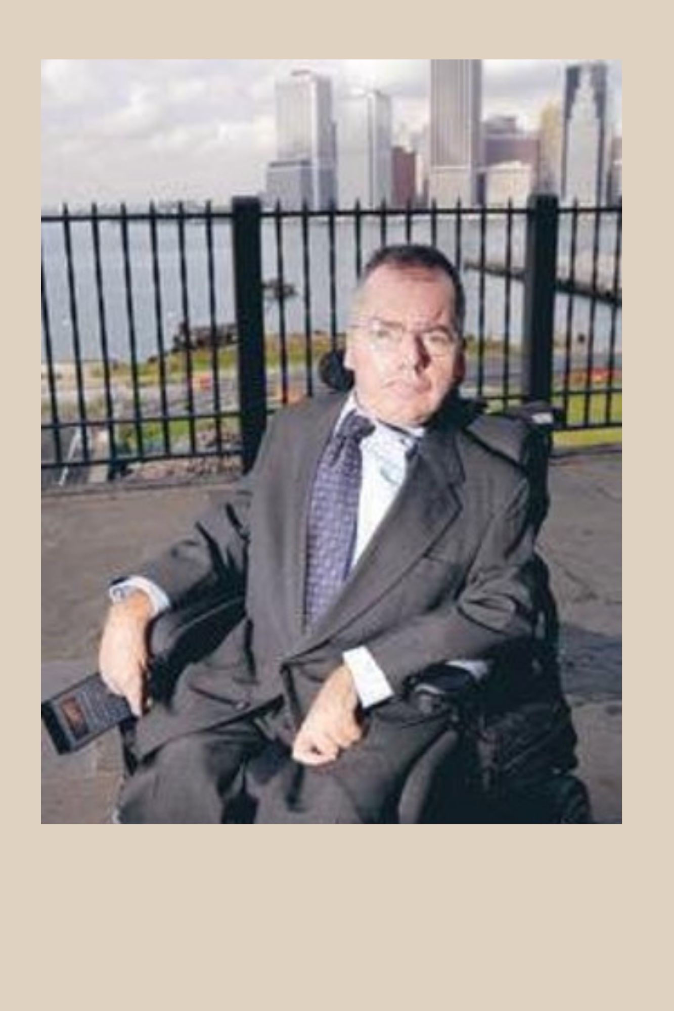 Photo of T.K. Small. He is wearing a suit. He is sitting in a wheelchair with a backdrop against a large city.