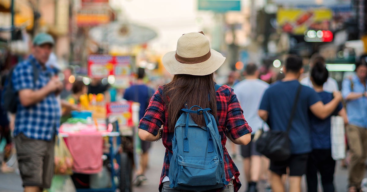 Solo female traveler in crowded urban market street – wearing backpack and hat, potential target for tourist scams