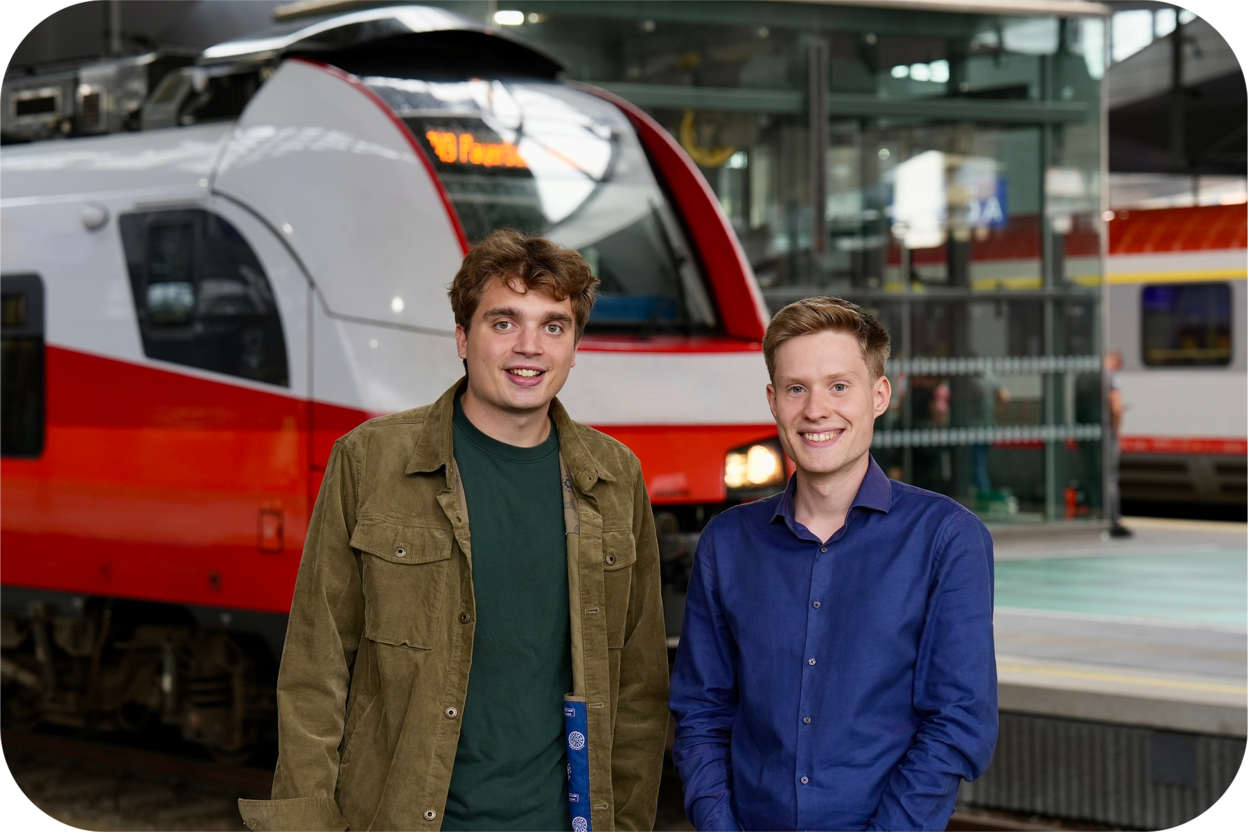 Zwei junge Männer stehen vor einem Zug und lächeln in die Kamera.