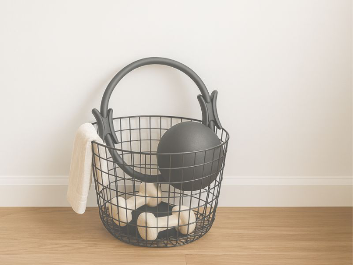 Pilates props in a black wire basket with ring, small ball, and hand weights.