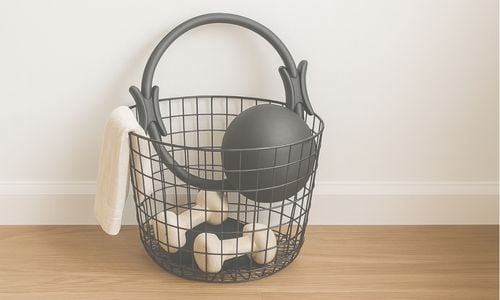 Pilates props in a black wire basket, including ring, small exercise ball, and hand weights